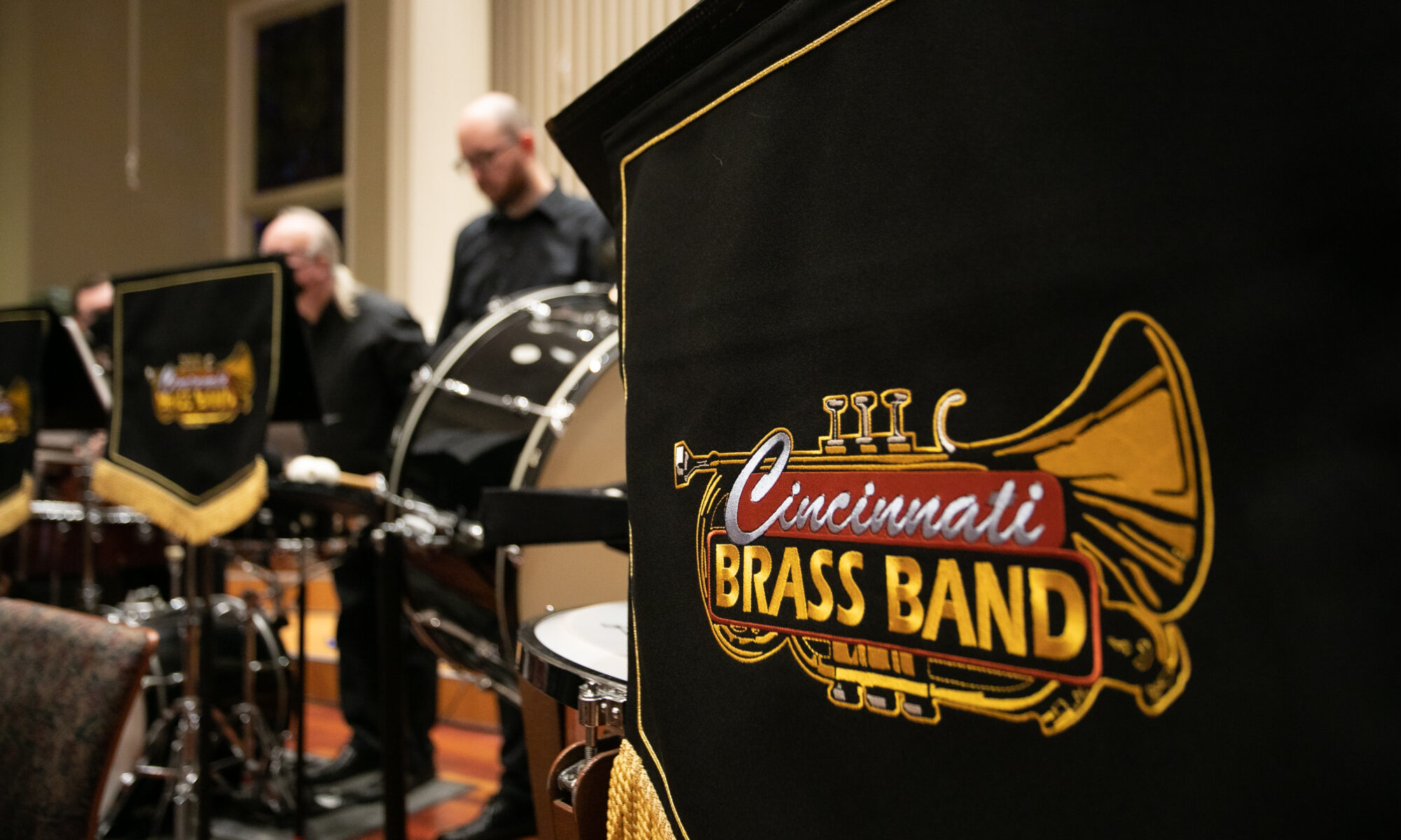 A music stand banner with the Cincinnati Brass band logo in the foreground and some of our percussion players in the background